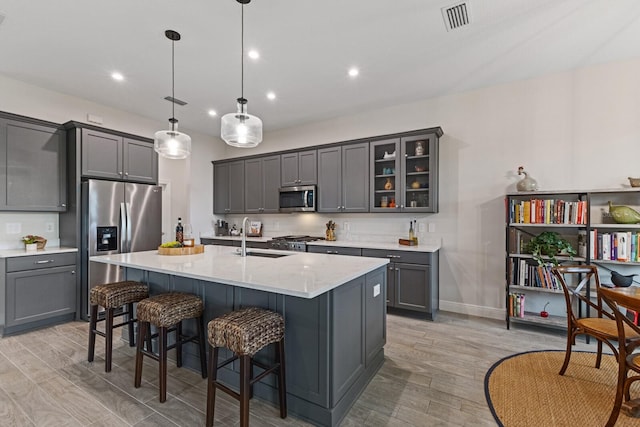 kitchen with a center island with sink, stainless steel appliances, gray cabinetry, hanging light fixtures, and sink