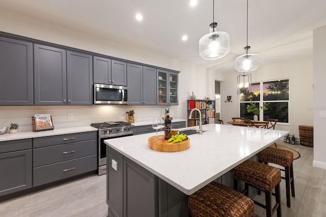 kitchen featuring light hardwood / wood-style floors, a center island with sink, gray cabinets, appliances with stainless steel finishes, and hanging light fixtures