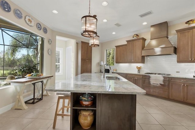 kitchen featuring sink, premium range hood, decorative light fixtures, tasteful backsplash, and a kitchen island with sink