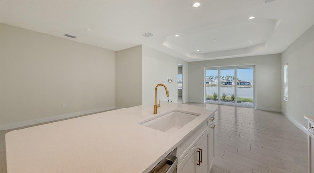 kitchen with a raised ceiling, a kitchen island with sink, a water view, white cabinets, and sink