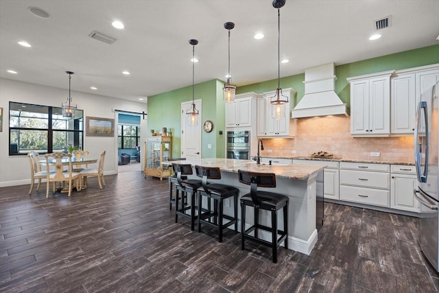 kitchen with premium range hood, white cabinetry, a kitchen island with sink, and hanging light fixtures