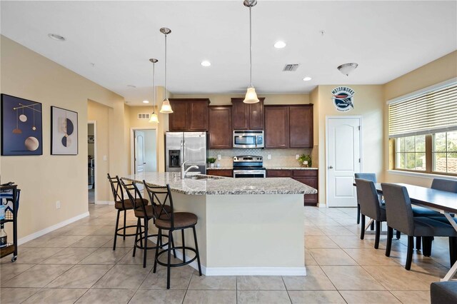 kitchen with a center island with sink, sink, hanging light fixtures, appliances with stainless steel finishes, and light stone counters