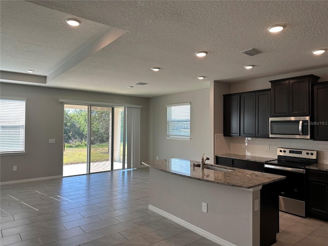 kitchen with an island with sink, appliances with stainless steel finishes, decorative backsplash, light stone counters, and sink