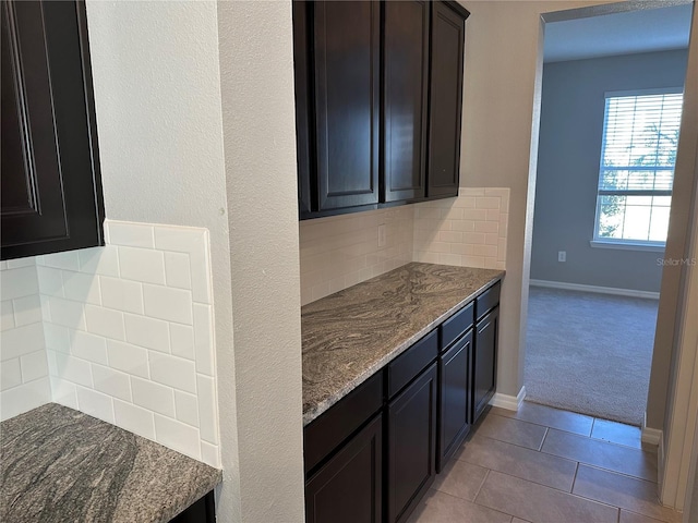 kitchen with light colored carpet, dark brown cabinetry, dark stone counters, and tasteful backsplash