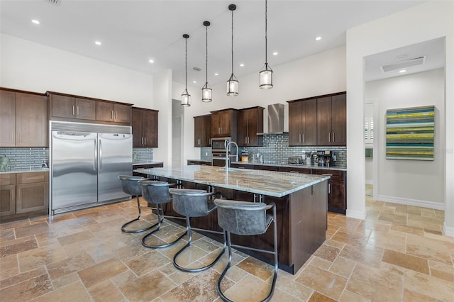 kitchen with hanging light fixtures, stainless steel appliances, light stone counters, an island with sink, and wall chimney exhaust hood