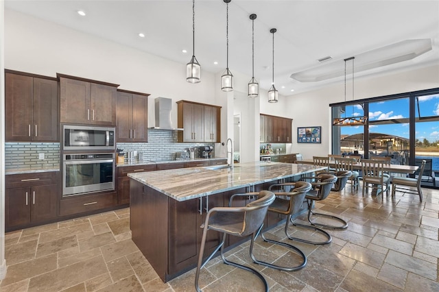 kitchen with wall chimney exhaust hood, light stone counters, hanging light fixtures, a center island with sink, and stainless steel appliances