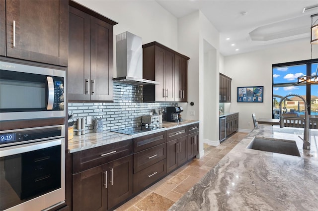 kitchen featuring sink, hanging light fixtures, stainless steel appliances, light stone countertops, and wall chimney range hood