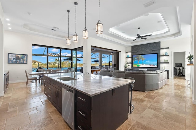 kitchen with a raised ceiling, hanging light fixtures, a kitchen island with sink, and stainless steel dishwasher