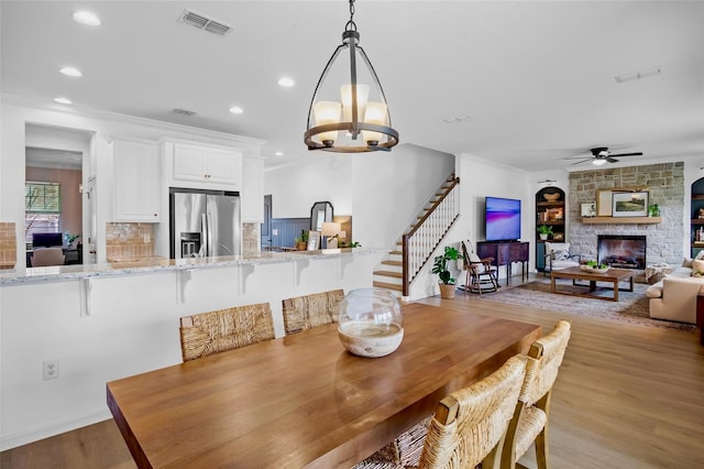 dining space with ceiling fan with notable chandelier, light hardwood / wood-style flooring, ornamental molding, and a stone fireplace