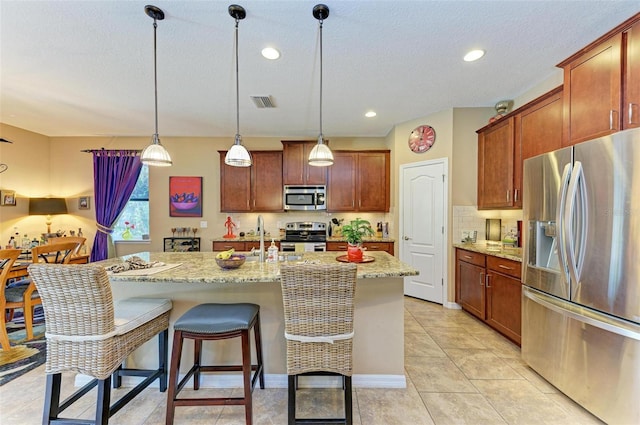 kitchen featuring pendant lighting, light tile patterned floors, appliances with stainless steel finishes, a kitchen island with sink, and light stone countertops