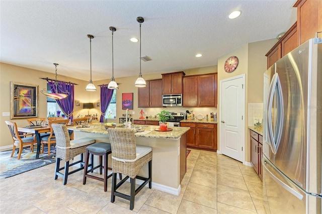 kitchen featuring appliances with stainless steel finishes, hanging light fixtures, a kitchen breakfast bar, light stone countertops, and an island with sink