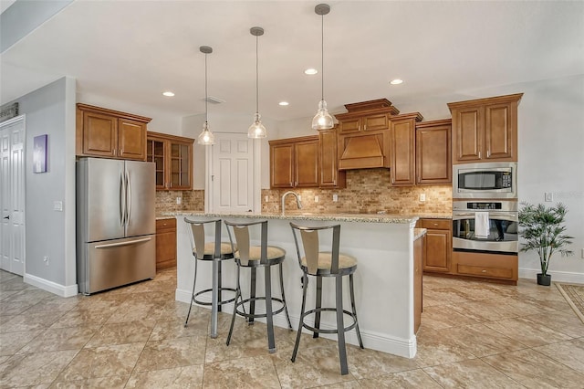 kitchen with appliances with stainless steel finishes, hanging light fixtures, a kitchen island with sink, light stone countertops, and wall chimney exhaust hood