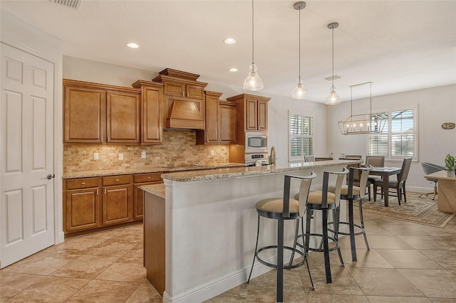 kitchen with a kitchen island with sink, stainless steel appliances, light stone counters, custom range hood, and decorative light fixtures