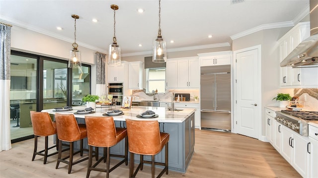 kitchen with appliances with stainless steel finishes, wall chimney exhaust hood, decorative light fixtures, white cabinetry, and a kitchen island with sink