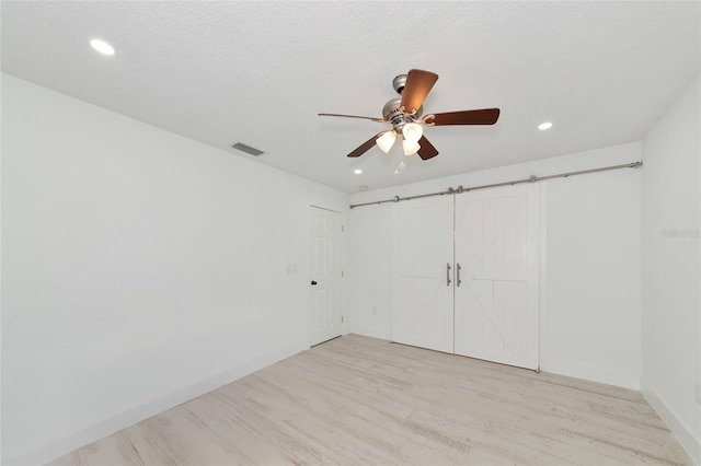 unfurnished bedroom with ceiling fan, light hardwood / wood-style floors, a barn door, a textured ceiling, and a closet