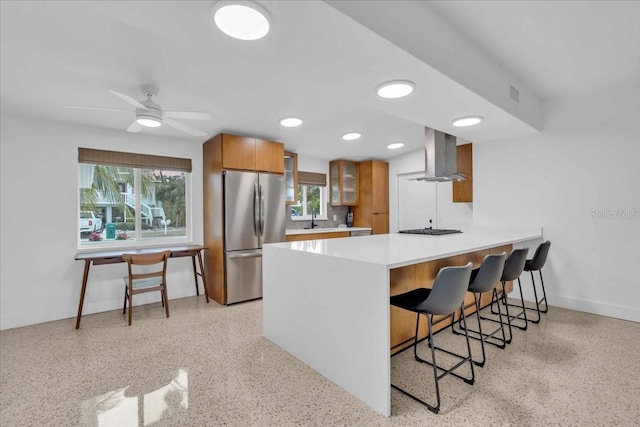 kitchen with ceiling fan, wall chimney range hood, kitchen peninsula, a kitchen bar, and stainless steel refrigerator