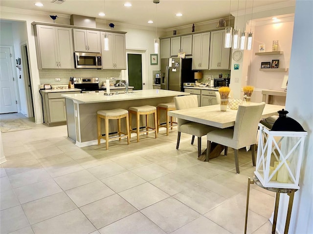 kitchen with gray cabinets, appliances with stainless steel finishes, hanging light fixtures, an island with sink, and decorative backsplash