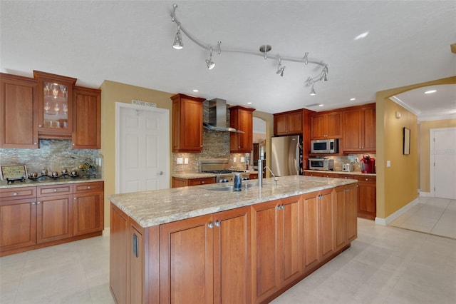 kitchen with light stone counters, appliances with stainless steel finishes, wall chimney exhaust hood, and a kitchen island with sink