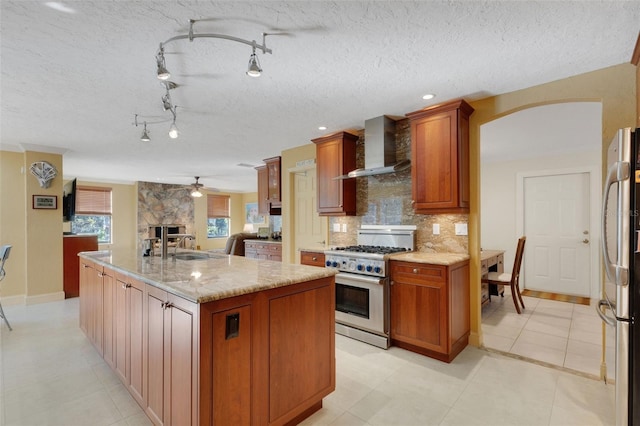 kitchen with a kitchen island with sink, backsplash, stainless steel appliances, light stone counters, and wall chimney exhaust hood