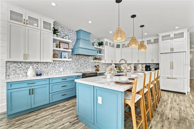 kitchen with white cabinetry, custom exhaust hood, a kitchen island with sink, decorative light fixtures, and blue cabinets