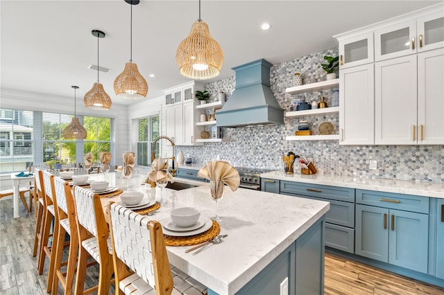 kitchen featuring custom exhaust hood, a kitchen island with sink, white cabinetry, and pendant lighting