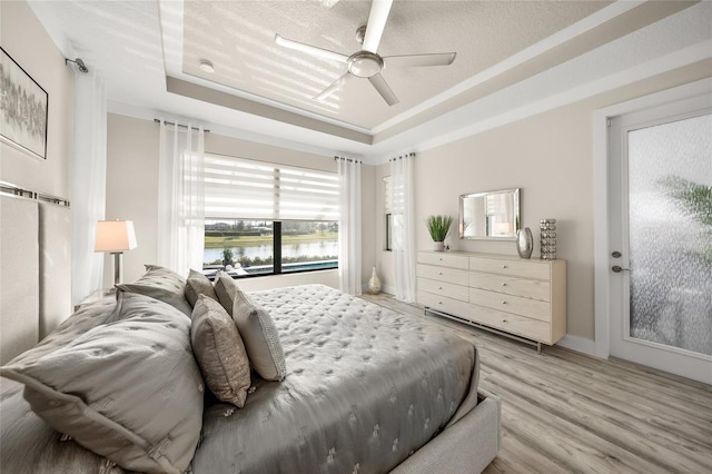 bedroom with a tray ceiling, light wood-type flooring, ceiling fan, and a water view