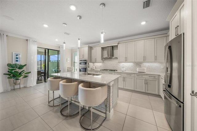 kitchen featuring pendant lighting, wall chimney range hood, sink, stainless steel appliances, and an island with sink