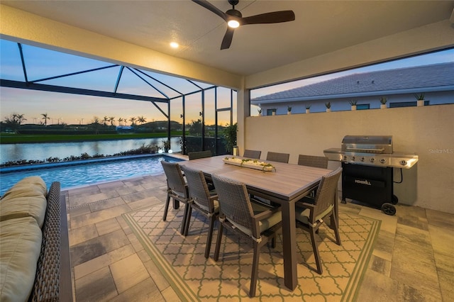 patio terrace at dusk featuring a water view, grilling area, glass enclosure, ceiling fan, and a fenced in pool