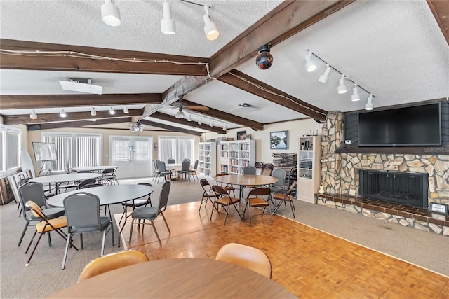 carpeted dining area with lofted ceiling with beams, a textured ceiling, a stone fireplace, and ceiling fan