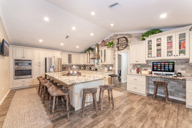 kitchen featuring stainless steel fridge with ice dispenser, light stone counters, wall chimney range hood, a large island with sink, and lofted ceiling