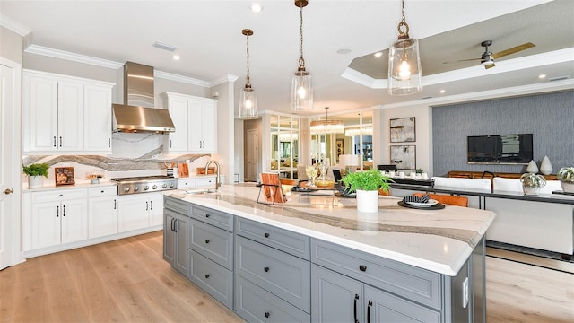 kitchen featuring white cabinetry, wall chimney range hood, hanging light fixtures, ceiling fan, and a center island with sink