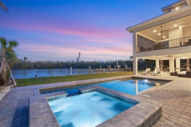 pool at dusk featuring a water view, a patio, ceiling fan, and an in ground hot tub