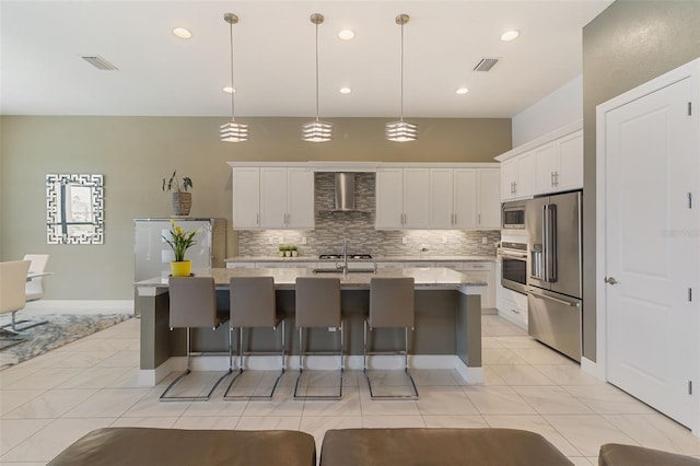 kitchen with pendant lighting, wall chimney range hood, stainless steel appliances, white cabinets, and a center island with sink