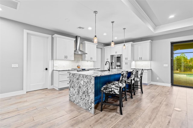 kitchen with white cabinetry, a center island with sink, appliances with stainless steel finishes, light stone countertops, and wall chimney range hood