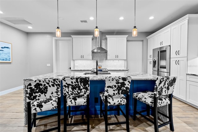 kitchen featuring stainless steel refrigerator with ice dispenser, a center island with sink, hanging light fixtures, and wall chimney range hood