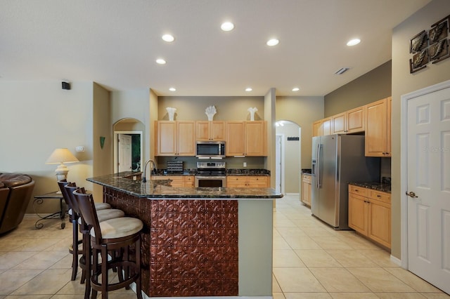 kitchen with stainless steel appliances, a kitchen bar, a kitchen island with sink, and dark stone counters