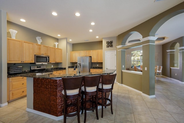 kitchen with light tile patterned flooring, light brown cabinetry, appliances with stainless steel finishes, an island with sink, and dark stone counters