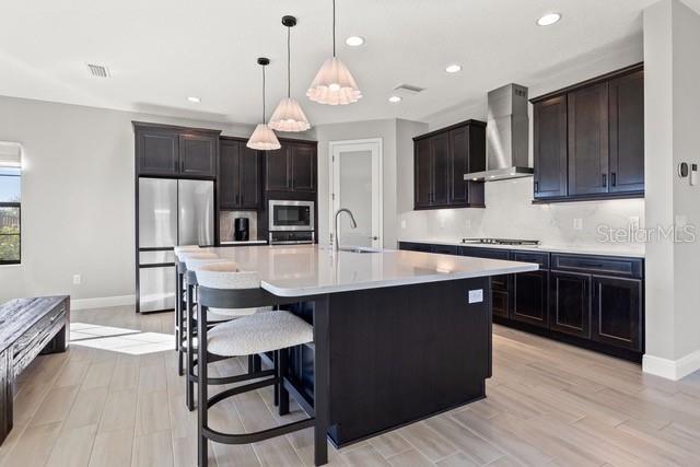 kitchen featuring decorative light fixtures, wall chimney range hood, stainless steel appliances, sink, and a center island with sink