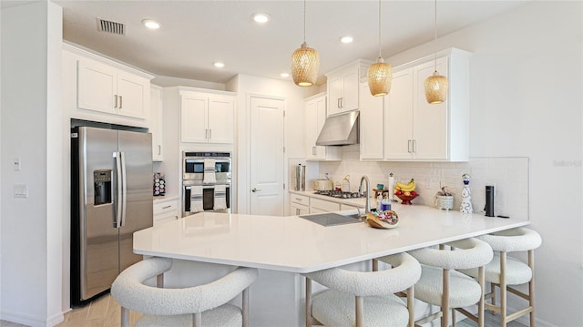 kitchen with under cabinet range hood, a peninsula, appliances with stainless steel finishes, backsplash, and a kitchen bar