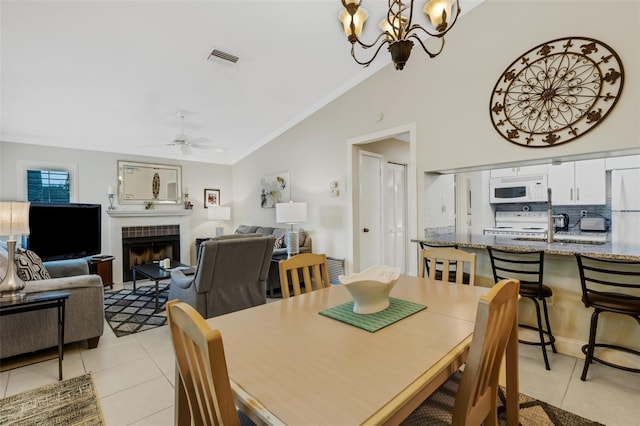 dining space with vaulted ceiling, ceiling fan with notable chandelier, a tiled fireplace, and light tile patterned flooring