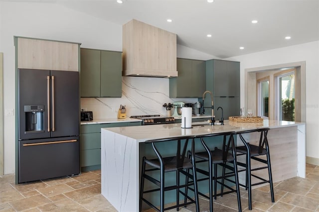 kitchen featuring a breakfast bar, high end black refrigerator, lofted ceiling, a kitchen island with sink, and light stone counters