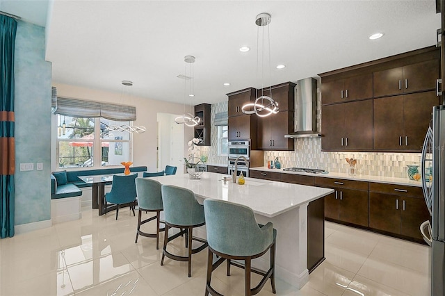 kitchen featuring hanging light fixtures, wall chimney range hood, an island with sink, and light countertops