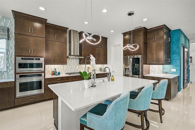 kitchen with decorative light fixtures, stainless steel appliances, backsplash, a kitchen island with sink, and wall chimney range hood