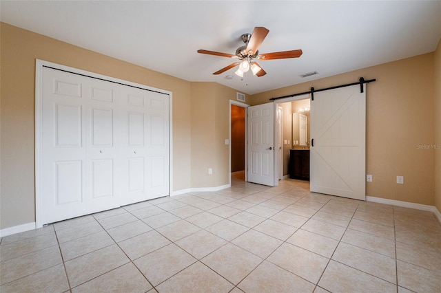 unfurnished bedroom featuring ensuite bathroom, a barn door, a closet, ceiling fan, and light tile patterned flooring