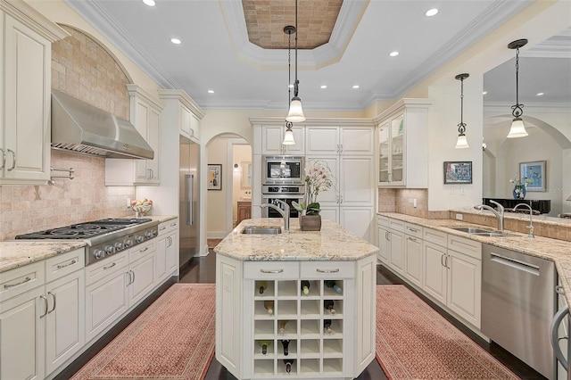 kitchen featuring built in appliances, decorative light fixtures, a tray ceiling, an island with sink, and wall chimney range hood