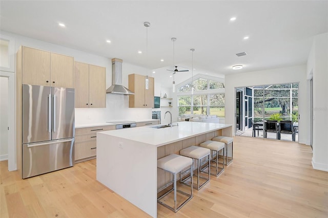 kitchen with decorative light fixtures, stainless steel fridge, light brown cabinets, a kitchen island with sink, and wall chimney range hood