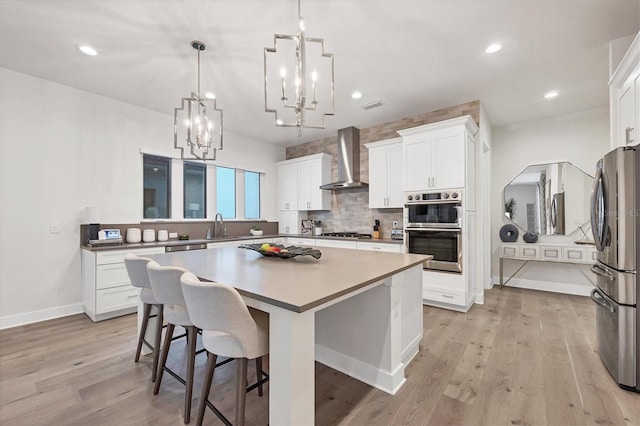 kitchen with wall chimney range hood, appliances with stainless steel finishes, a center island, white cabinets, and decorative light fixtures