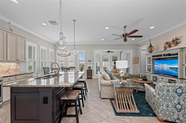 kitchen with sink, tasteful backsplash, crown molding, hanging light fixtures, and an island with sink