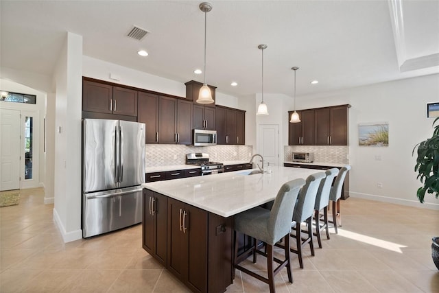 kitchen featuring a kitchen island with sink, hanging light fixtures, dark brown cabinets, stainless steel appliances, and tasteful backsplash