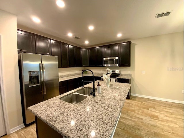 kitchen featuring dark brown cabinetry, sink, appliances with stainless steel finishes, an island with sink, and light stone countertops